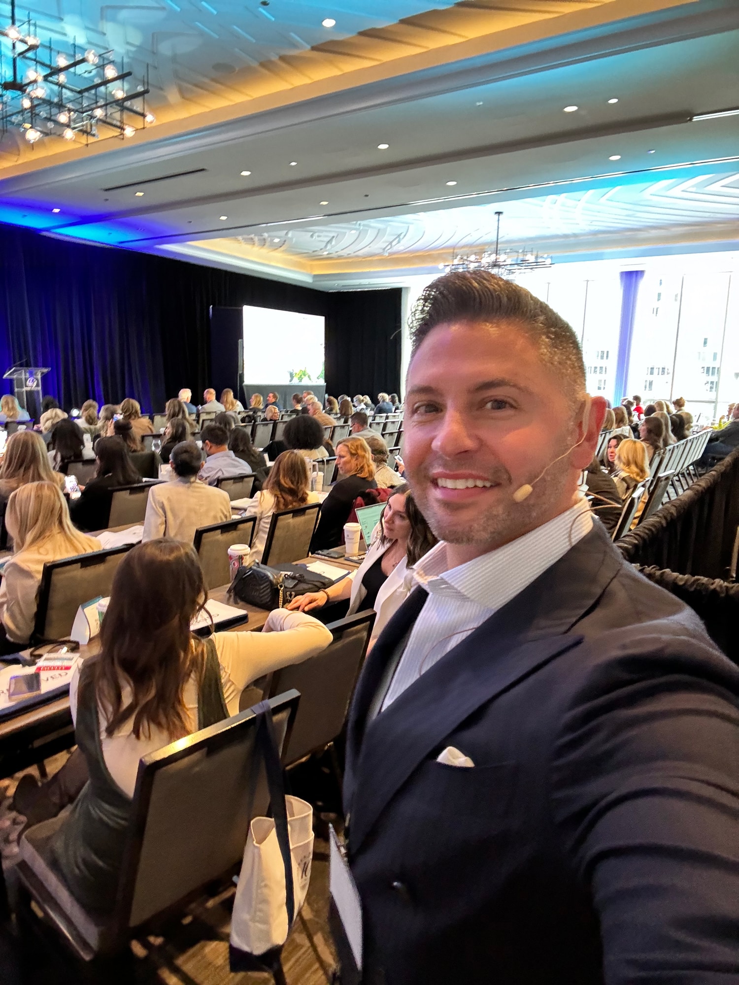 A man in a suit takes a selfie at a conference with an audience in the background.