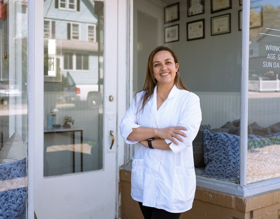 A smiling woman in a lab coat stands outside a clinic, arms crossed, with certificates on the wall behind her.