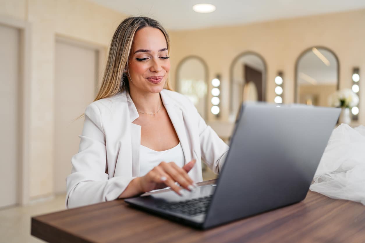 A woman in a white blazer using a laptop at a wooden desk indoors.