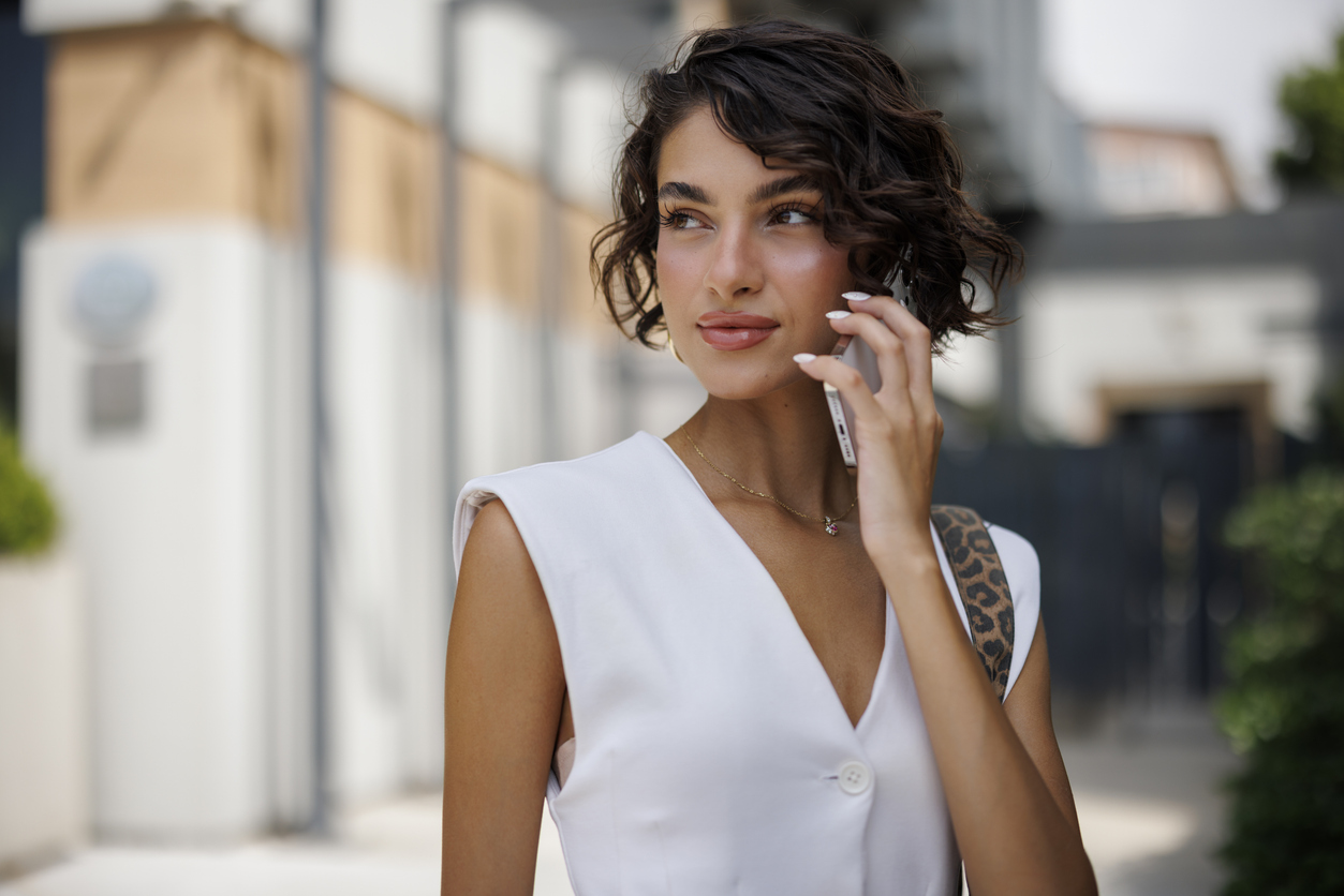 A woman in a white dress talks on a smartphone outdoors.